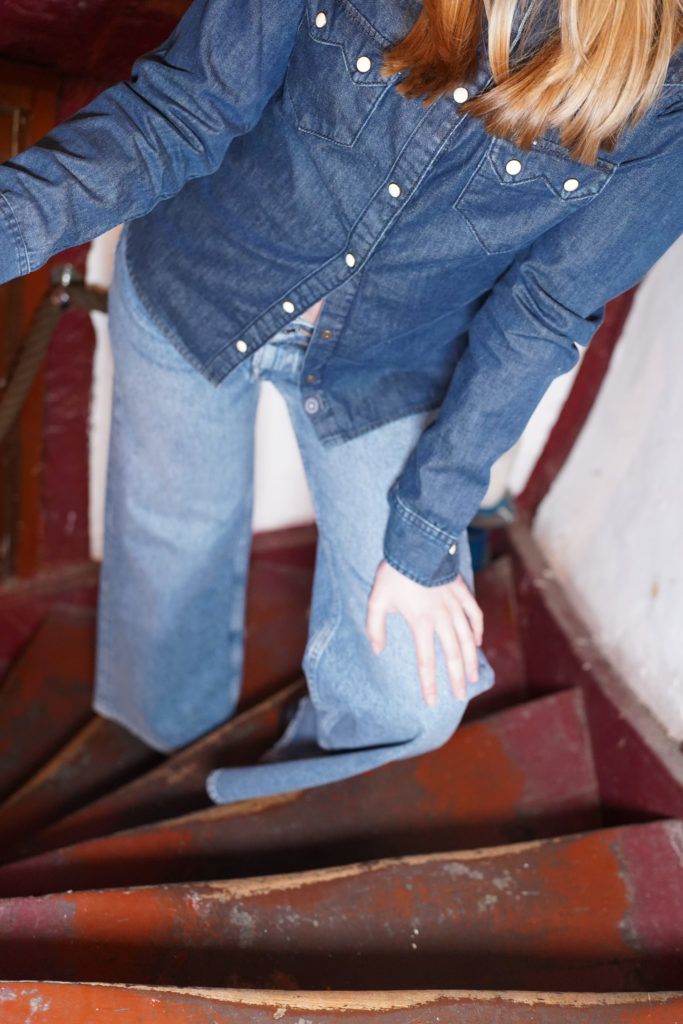 Janine in Kuyichi Sawtooth Shirt Worker Blue auf der Treppenmitte (nur das Shirt und die Hose sind zu sehen)